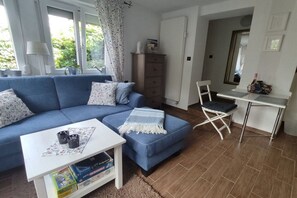 A living room with a blue sofa, a white coffee table, and a small dining area with a white chair and table. - Holiday apartment with 2 bedrooms (Wilhelmshaven)