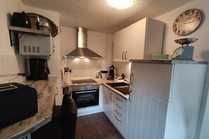 Ett kompakt kök med vita skåp, en rostfri köksfläkt, en inbyggd ugn, en kaffebryggare och en antik klocka.