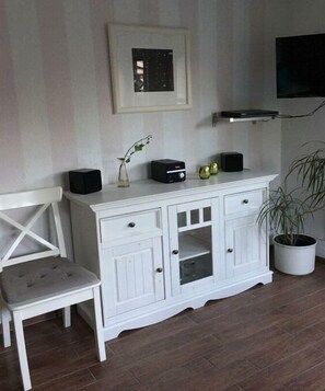 A white cabinet with drawers and a glass door, a small table with a vase and a plant, and a television mounted on the wall. - Holiday apartment with 2 bedrooms (Wilhelmshaven)