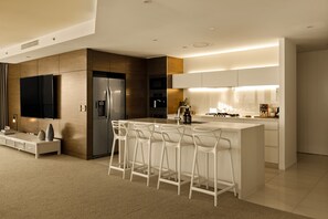 A modern kitchen with a central island, bar stools, a built-in oven, and a flat-screen TV mounted on the wall. - Two Horizons by Aryve (Broadbeach)