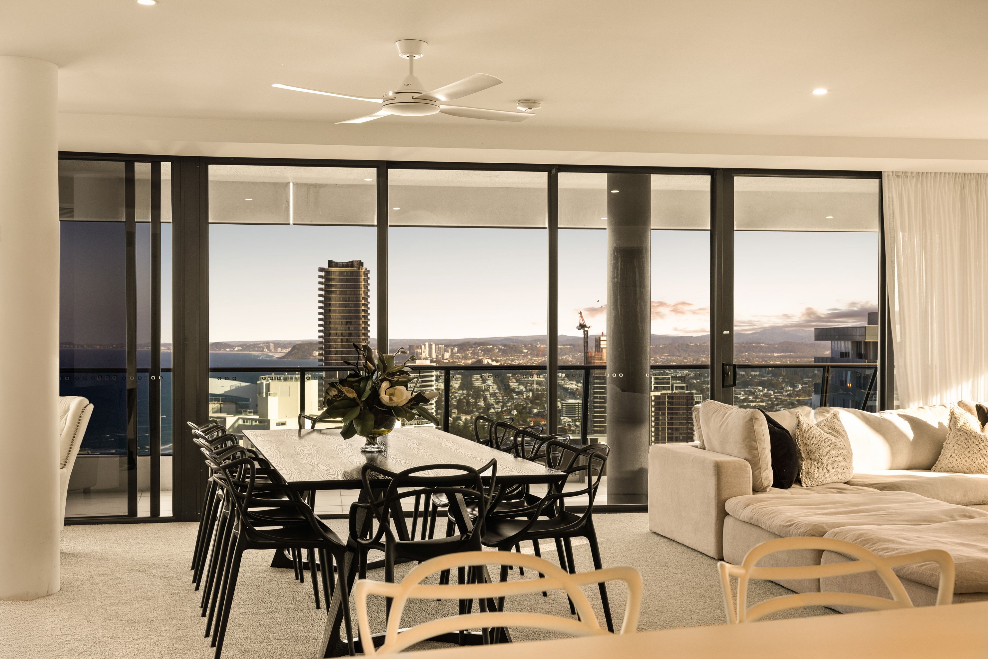 A modern dining area with a large table and chairs, a ceiling fan, and a view of a cityscape through floor-to-ceiling windows.