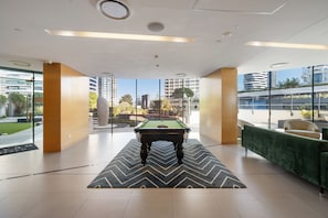 A pool table in a modern indoor space with large windows, a green sofa, and a view of a cityscape. - Two Horizons by Aryve (Broadbeach)