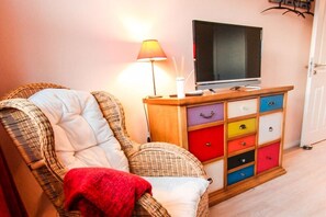 A wicker chair with a cushion and a red blanket, a television on a wooden cabinet with colorful drawers, and a lamp with a white shade. - Holiday home with 2 bedrooms (Fuhlendorf)