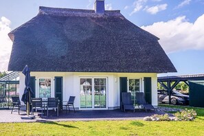 A house with a thatched roof, a covered patio with chairs, and a car parked in the background. - Holiday home with 2 bedrooms (Fuhlendorf)
