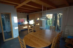 A dining area with a round wooden table and chairs, a kitchen in the background, and a view of the outdoors. - Holiday home with 2 bedrooms (Ostseebad Prerow)