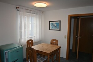 Ein Zimmer mit Holztisch und -stühlen, einem kleinen Kühlschrank, einem Fenster mit Vorhängen, einem gerahmten Bild an der Wand und einer Tür.