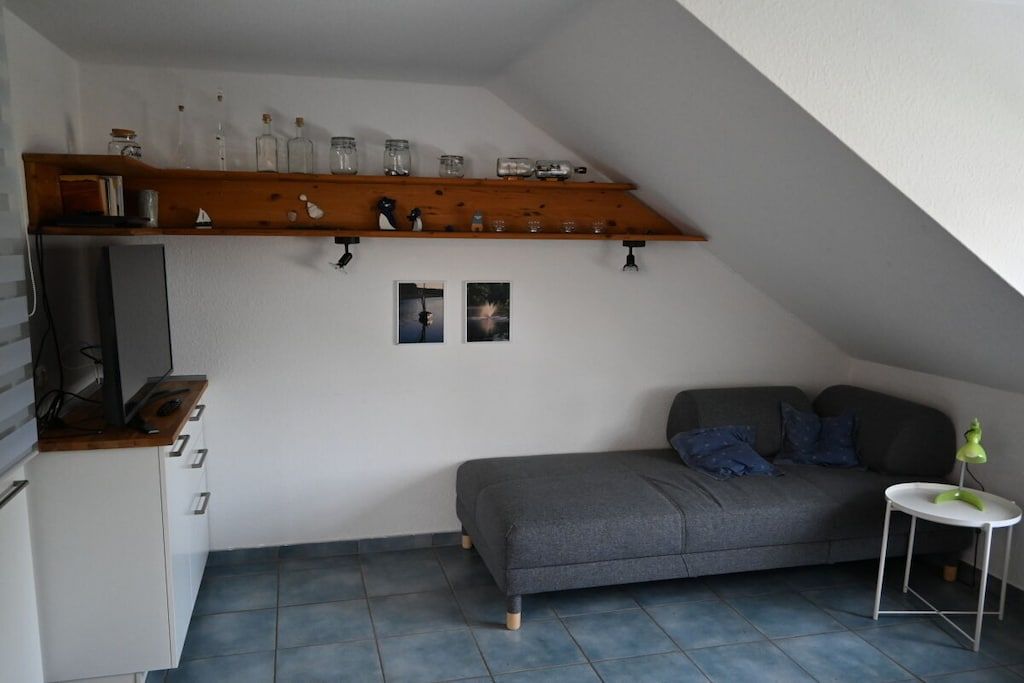 Een compacte, moderne woonruimte met een grijze bank, een houten plank met glazen potten, een tafeltje met een lamp en een televisie op een kast.