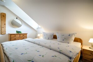 A bedroom with a sloped ceiling, a wooden nightstand, a bedside table, a mirror, and a bed with floral bedding. - Holiday home with 3 bedrooms (Ostseebad Prerow)
