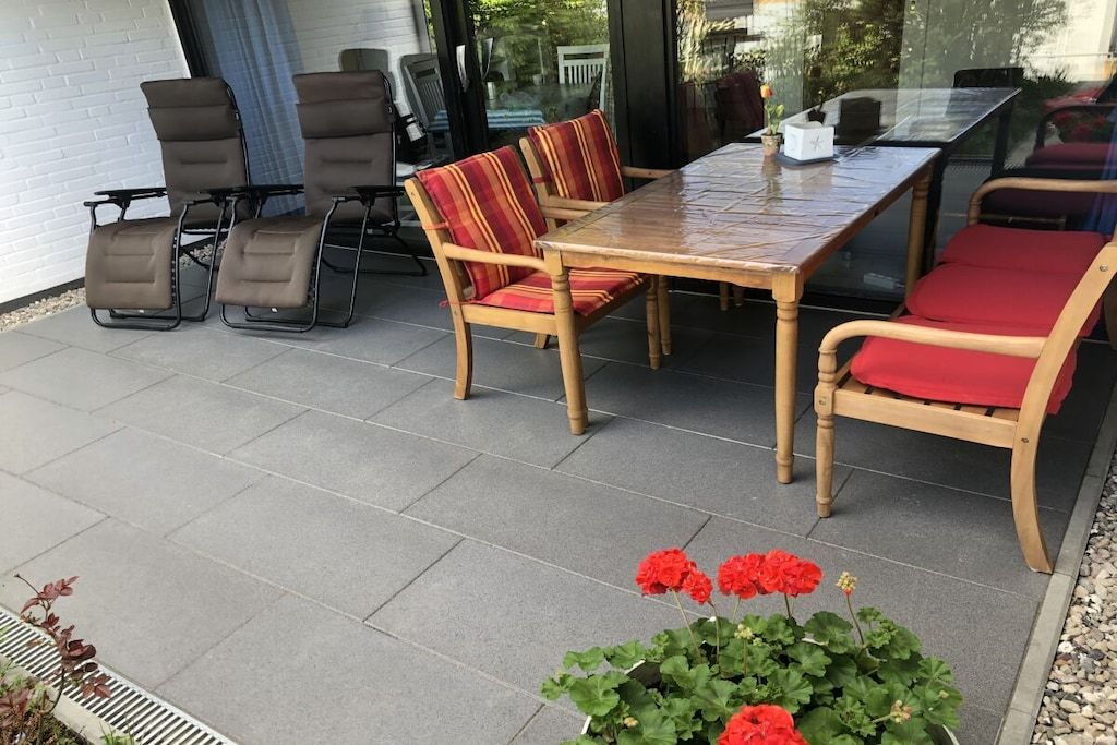 An outdoor patio with a wooden table and chairs, a glass partition, and a potted plant with red flowers.