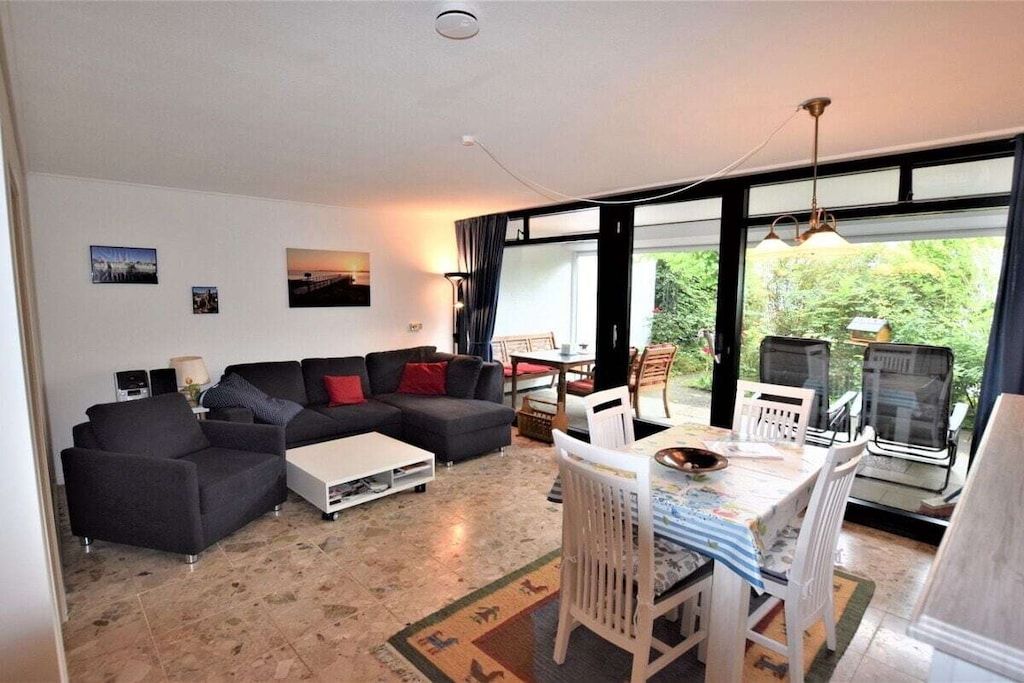 A living room with a sofa, coffee table, and dining area with chairs. There are windows and a view of greenery outside.