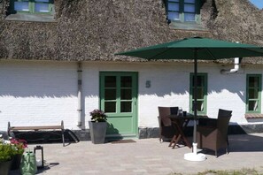 A thatched-roof building with a white exterior and green shutters. There is a patio with a table and chairs, a green umbrella, and potted plants. - Holiday home in Schlei with 2 bedrooms (Klappholz)