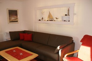 A living room with a brown sofa, a red chair, a small table with two red placemats, a framed picture on the wall, and a shelf with a model sailboat and decorative items. - Holiday home in Born with swimming pool (Born)