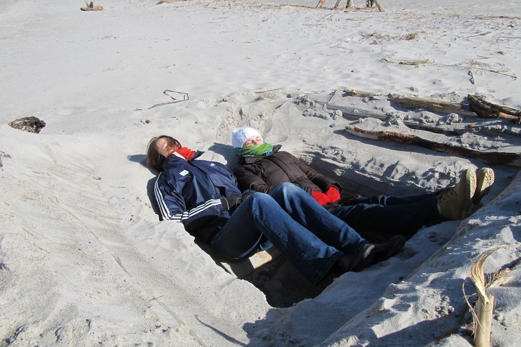 Dos personas descansando sobre arena, con una estructura de madera cerca.