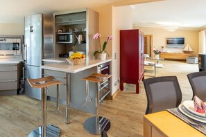 A modern kitchen with a stainless steel refrigerator, a microwave, and a fruit basket on the counter. There are two bar stools and a wooden dining table with chairs. - Penthouse Villa Gudrun with private sauna (Ostseebad Binz)