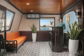 A reception area with a wooden ceiling, a sofa, a potted plant, a reception desk, and a view of a body of water through a window. - Halong Arcadia Cruise (Ha Long)