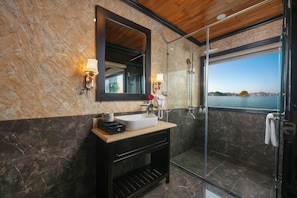 A bathroom with a marble wall, a large mirror, a sink, a shower, and a view of the sea. - Halong Arcadia Cruise (Ha Long)