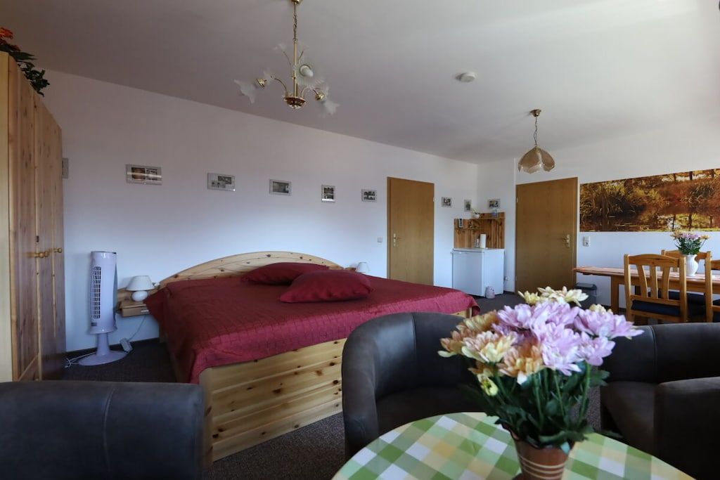 Ein Zimmer mit einem Bett, einem Tisch mit Blumen, einem Sessel, einem Ventilator und einem Bild an der Wand.