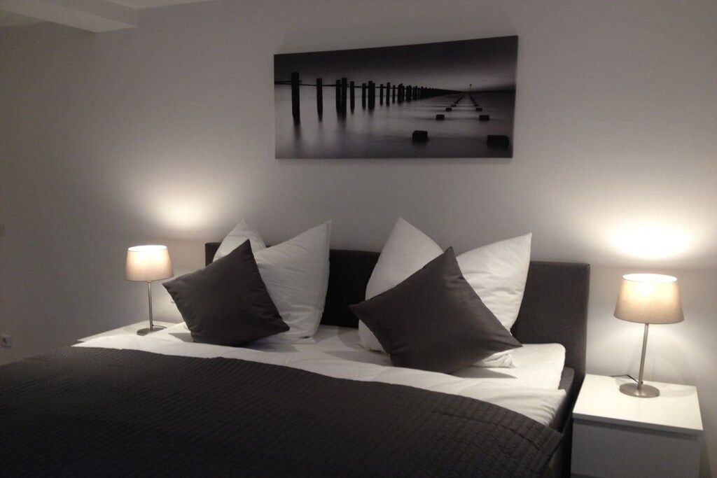 Una cama bien hecha con un cabecero oscuro y ropa de cama blanca, flanqueada por dos lámparas de noche en mesitas blancas.