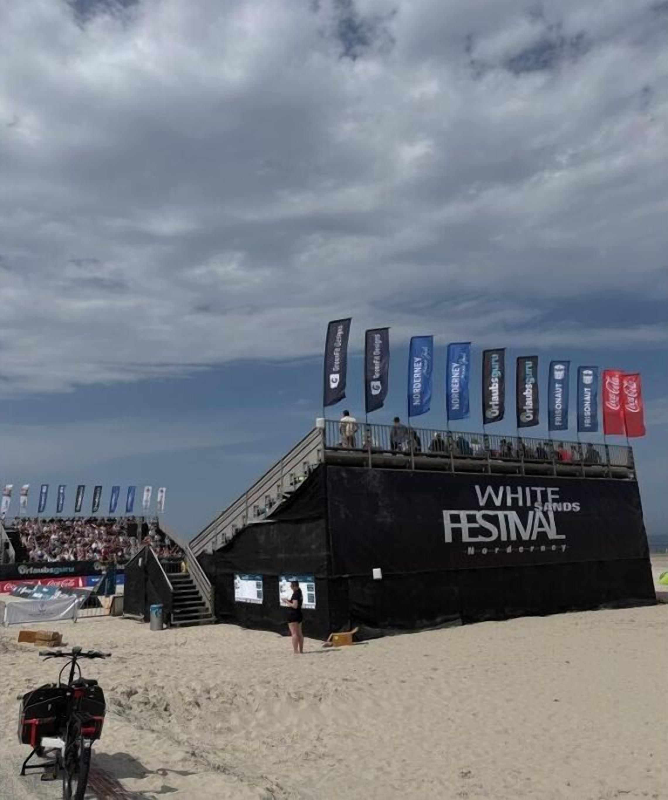 Un escenario con un cartel para el Festival de White Sands, un entorno playero y una bicicleta.