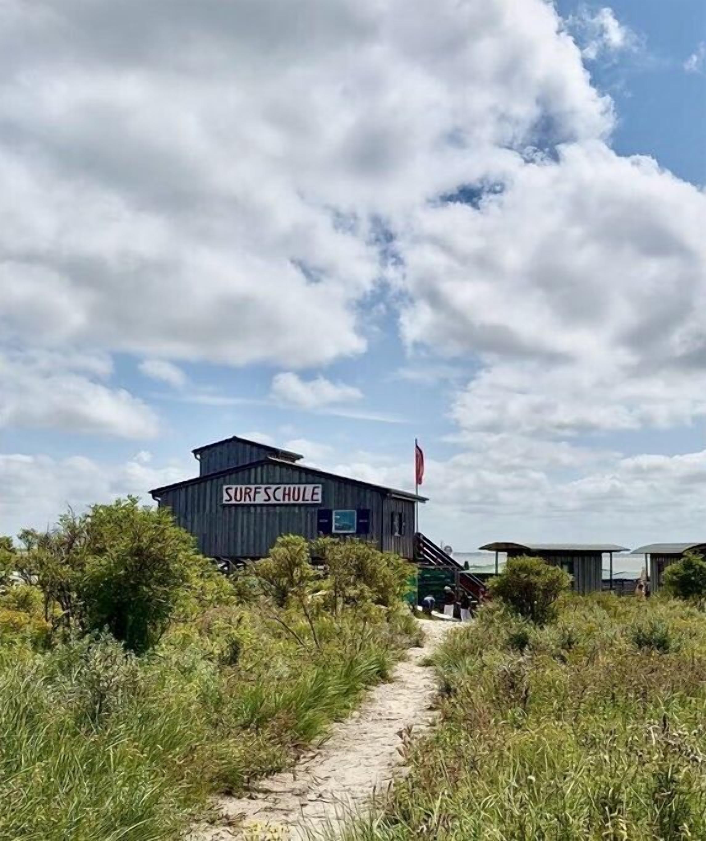 Un bâtiment portant l’inscription « SURFSCHULE », avec un chemin de sable menant à l’entrée, et entouré de végétation.