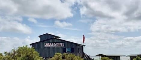 Un bâtiment portant l’inscription « SURFSCHULE », avec un chemin de sable menant à l’entrée, et entouré de végétation.
