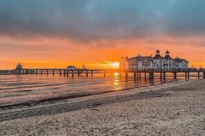 A coastal scene with a pier extending into the water, a large building with multiple stories, and a sunset in the background. - Holiday apartment KÜSTENNEST Baabe (Baabe)
