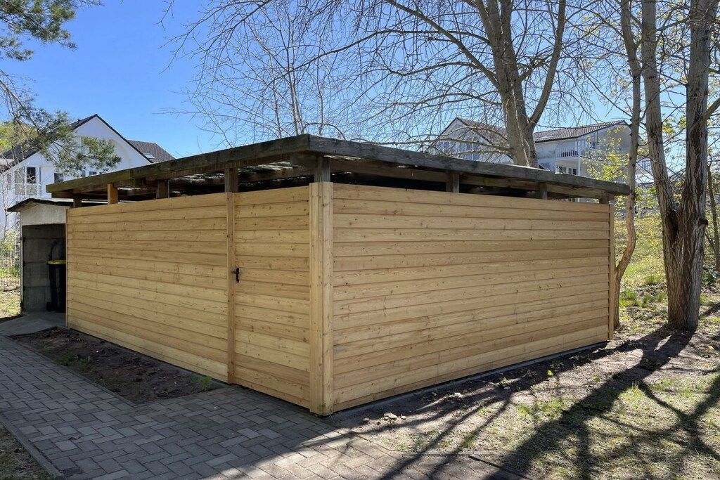 A wooden structure with a sloped roof, possibly a shed or storage unit, situated on a paved area with trees in the background.