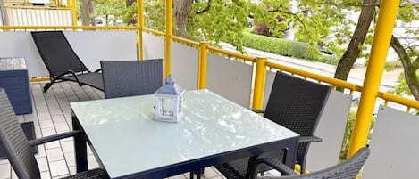 Un espace extérieur aménagé avec une table en verre et des chaises en osier, entouré de rampes jaunes et d’arbres.