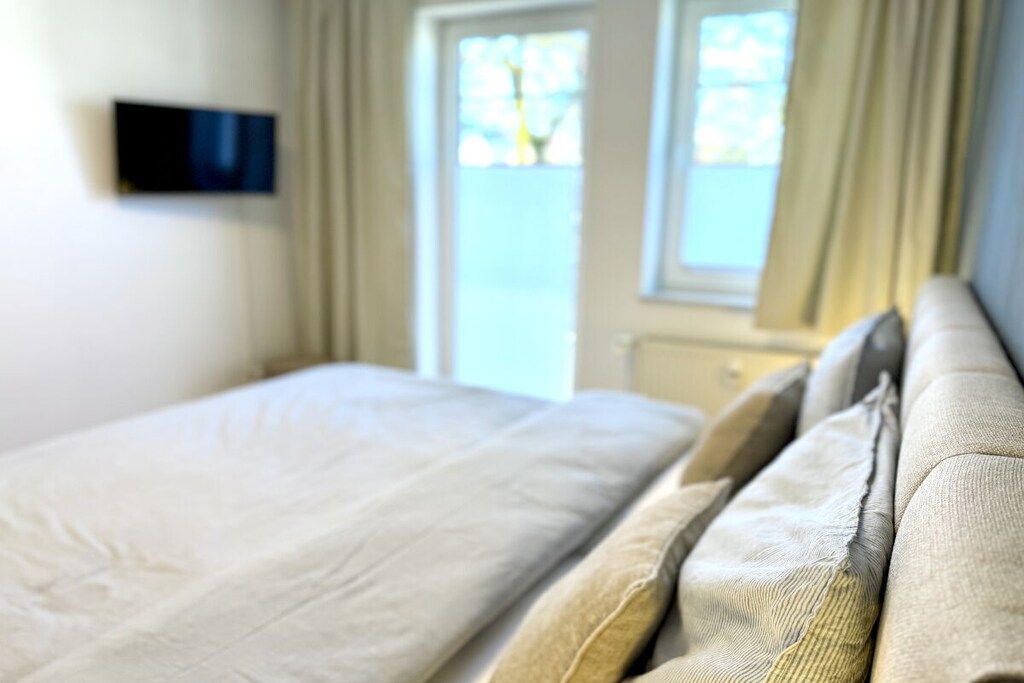 Ein Bett mit Kopfteil, ein an der Wand befestigter Fernseher und ein Fenster mit Vorhängen.
