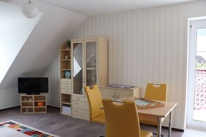 A room with a television, a cabinet with drawers, a table with chairs, and a window with blinds. - Holiday apartment in Süsel with swimming pool (Süsel)