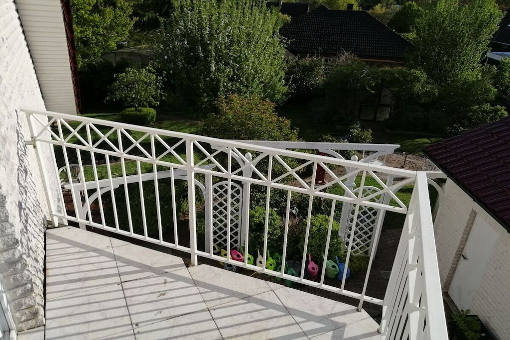 Ein Balkon mit weißer Brüstung, gefliestem Boden und Blick auf einen Garten mit Bäumen und einem Haus im Hintergrund.