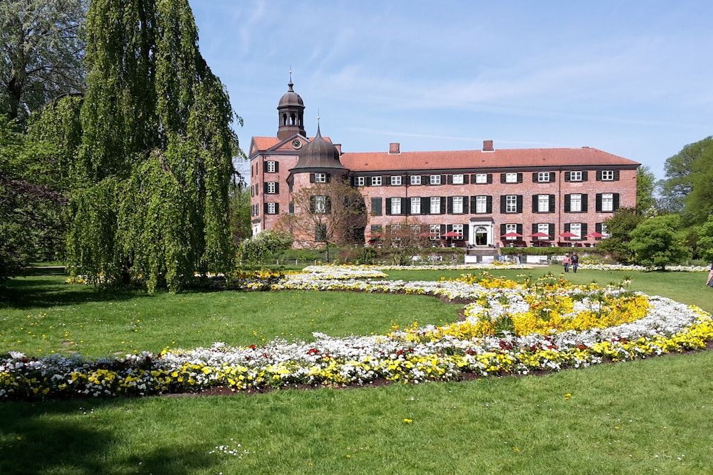 A large building with a tower, surrounded by a well-maintained garden with flower beds and a tall tree.