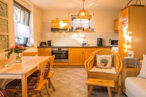 Une cuisine avec des meubles de rangement en bois, une table à manger avec des chaises, un luminaire suspendu et une fenêtre avec des rideaux.