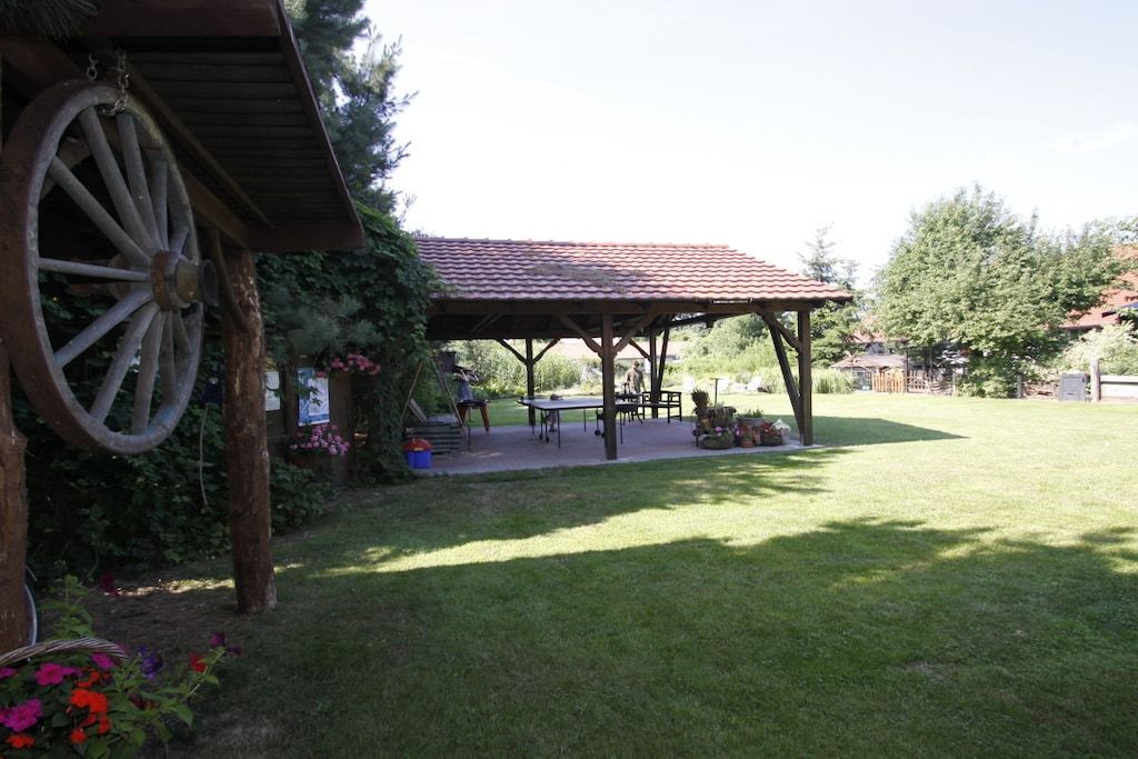 A covered outdoor area with a table and chairs, surrounded by a grassy lawn and trees.