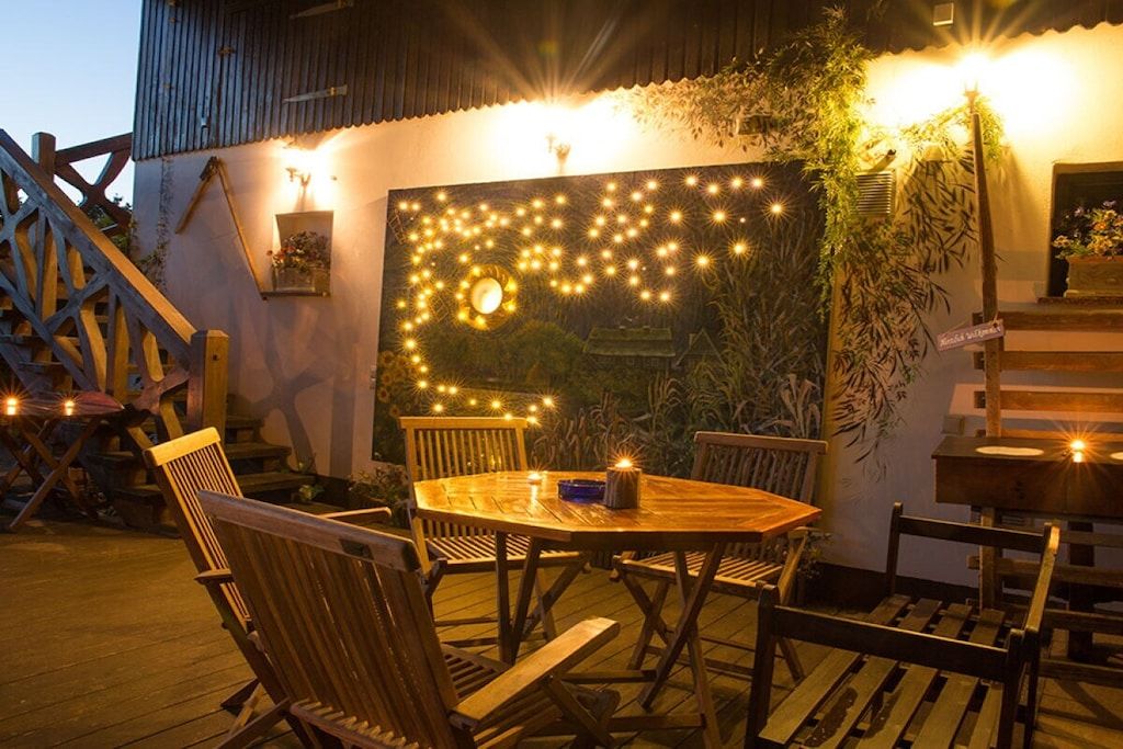 Un área de comedor al aire libre con una mesa y sillas de madera, rodeada por un muro decorado con luces y plantas.