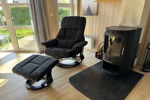 Ein Zimmer mit einem großen Fenster, einem schwarzen Sessel, einem schwarzen Hocker und einem schwarzen Kaminofen.