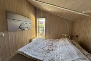 A bedroom with wooden walls, a bed with a white bedspread, a bedside table, a lamp, and a window. - Holiday home in the Baltic Sea with 2 bedrooms (Boiensdorf Stove)