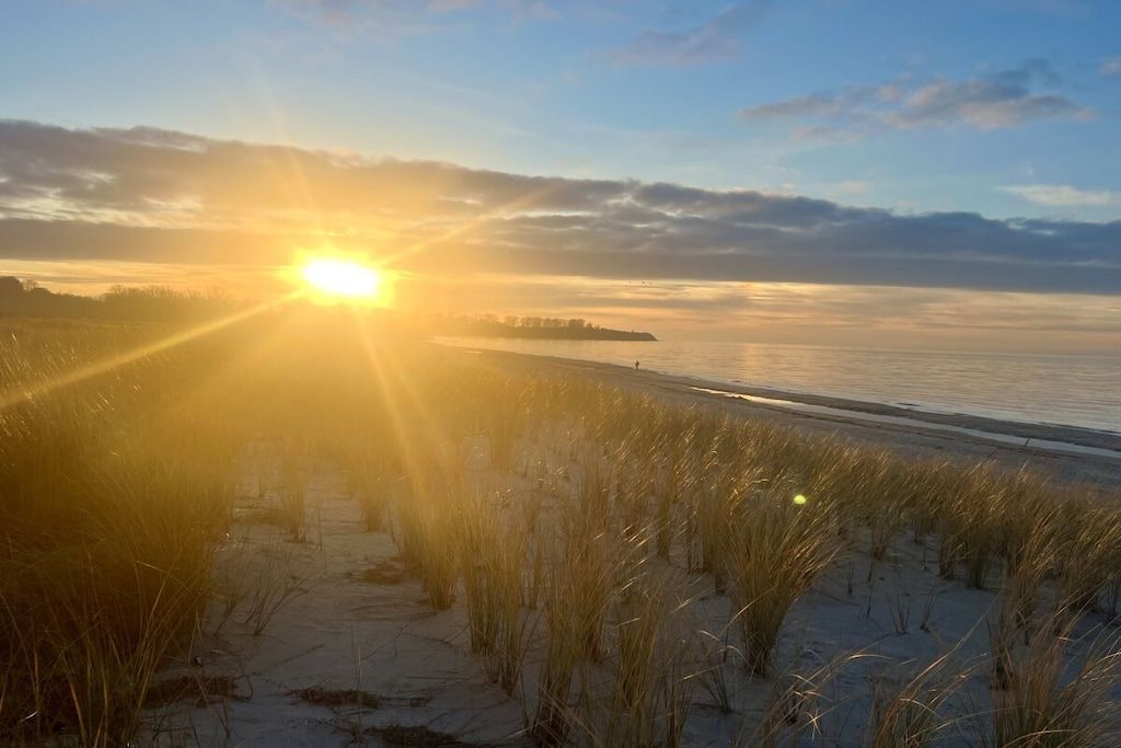 Ein Strand mit Sonnenuntergang, grasbewachsenen Dünen und einer fernen Küste.