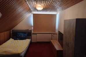 A small, single-bed room with a sloped ceiling, a radiator, a wooden desk, and a large window with blinds. - Bergner's Country House (Hollern-Twielenfleth)