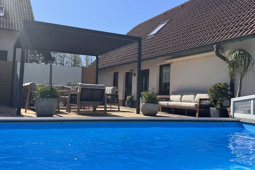 Una piscina con sillas de descanso y plantas en macetas, un patio cubierto y una casa con techo de tejas.