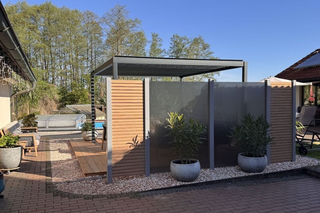 Un espacio de estar al aire libre cubierto con una terraza de madera, plantas en macetas y una parrilla.