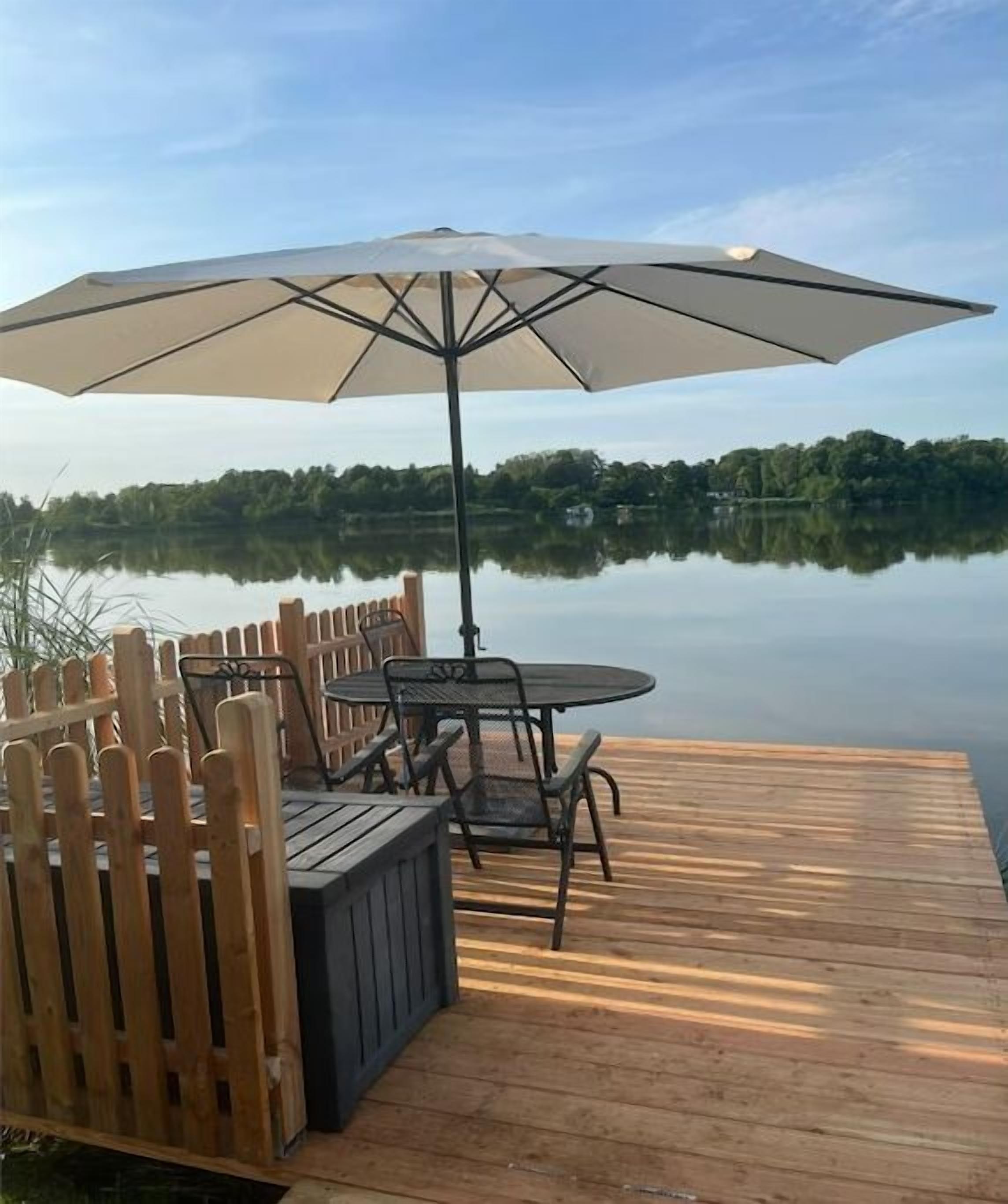A wooden deck with a table and chairs, an umbrella, and a view of a calm lake.