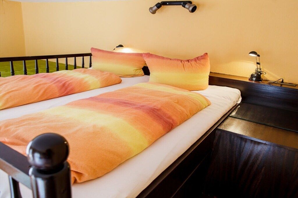A double bed with a headboard, two pillows, a bedside table with a lamp, and a wall-mounted light fixture.