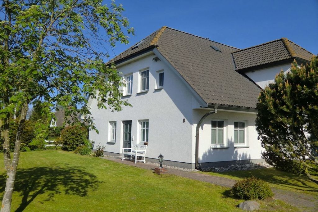 Une maison à deux étages, avec une façade blanche, un toit en tuiles et une pelouse devant, agrémentée d’un banc et d’un lampadaire.