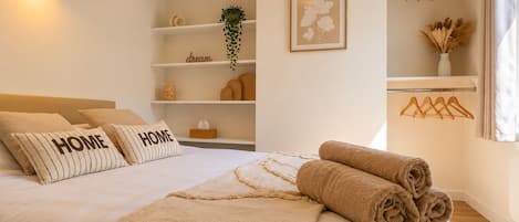 Un dormitorio con cama, almohadas, una mesita de noche, una lámpara colgante, un estante con adornos y una ventana con cortinas.