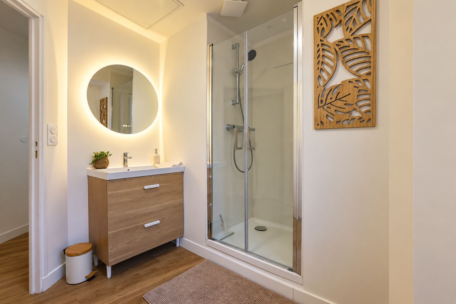 Un baño moderno con cabina de ducha de vidrio, tocador de madera con un espejo redondo y un panel decorativo en la pared.