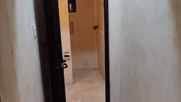 Un couloir étroit avec une porte donnant sur une pièce dont le sol est de couleur claire et qui contient une petite étagère sur laquelle sont posés divers objets.