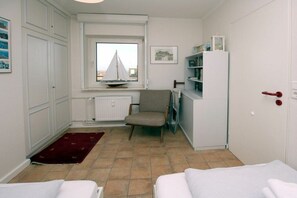 Ein kleines Zimmer mit Bett, Stuhl, Tisch, Fenster und einem Modell eines Segelboots sowie einer Heizung.