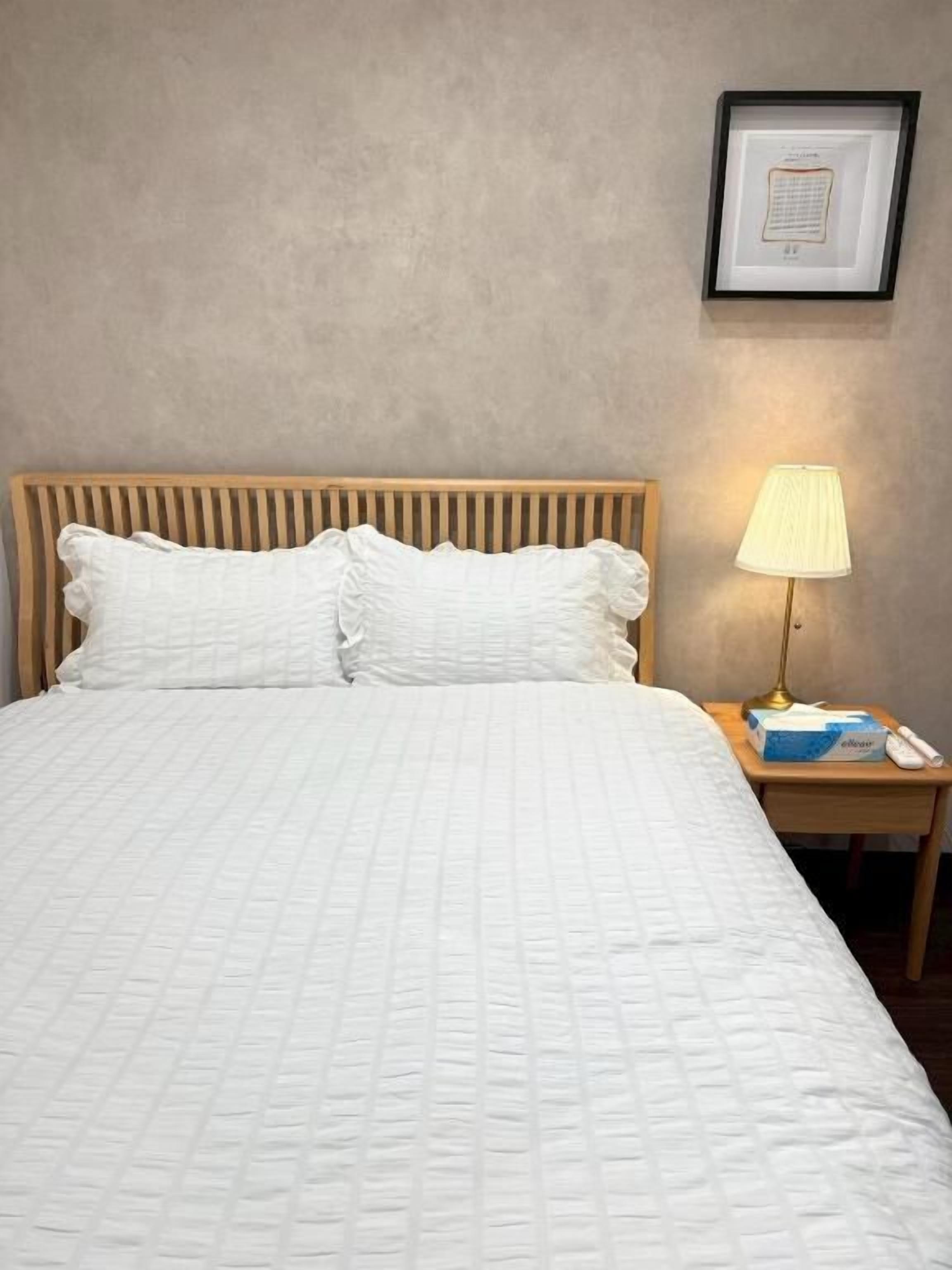 Una cama bien tendida con cabecera de madera y ropa de cama blanca. Hay una lámpara de noche y un cuadro en la pared.