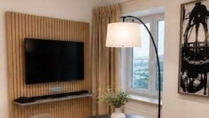 Una camera moderna con TV a schermo piatto fissata a un pannello di legno, un tavolo rotondo con due sedie, una lampada da terra e una finestra con tende.